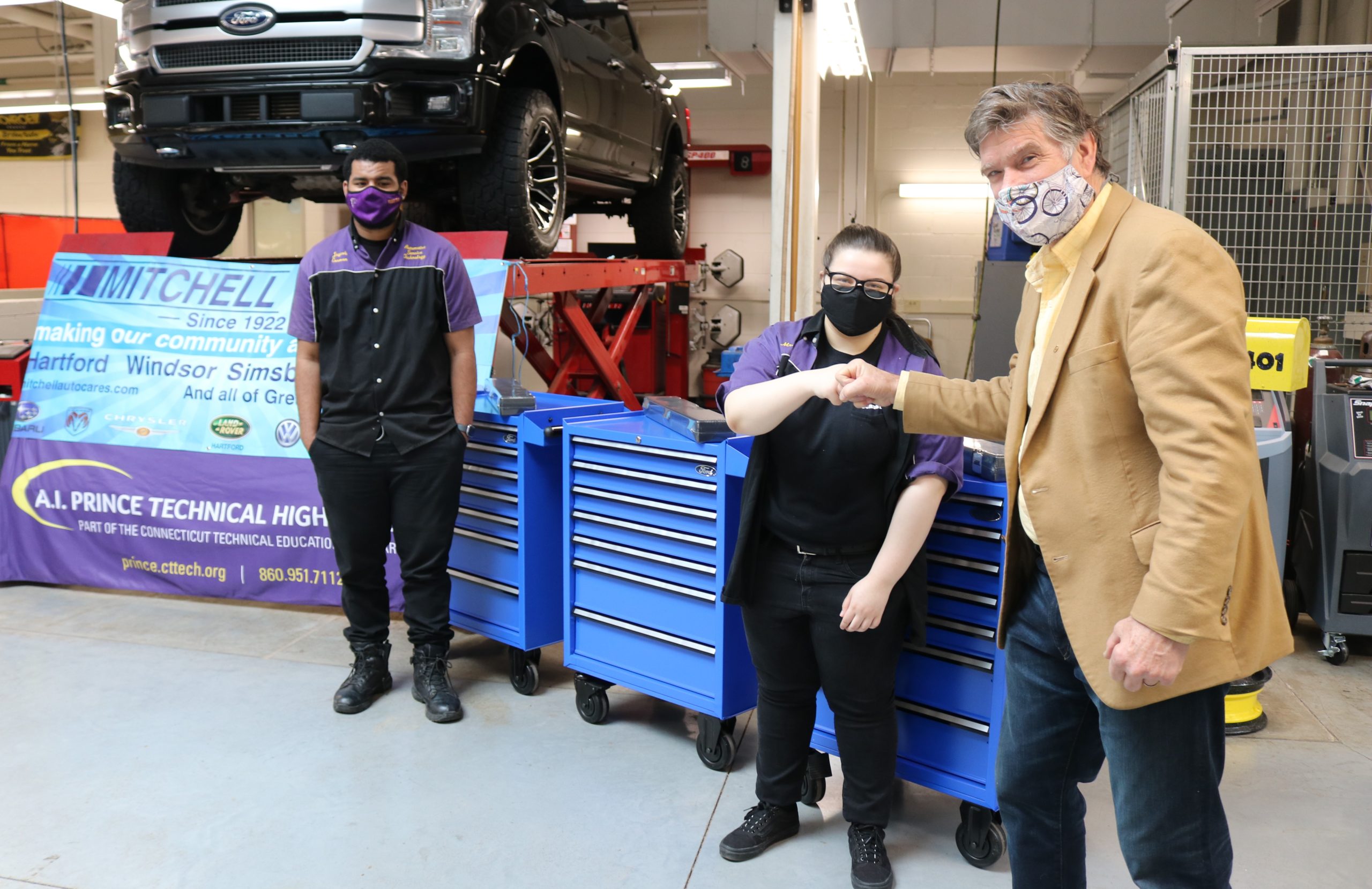 Steven Mitchell, Vice President of Mitchell Auto Group, stands next to two student recipients of the tool kits. Three blue tools kits are in the background, and a black pick up truck is on a car lift up in the air.i