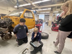 Two students sit on the floor with their Chromebook in front of a partially disassembled pick up truck and contemplate something on the screen.