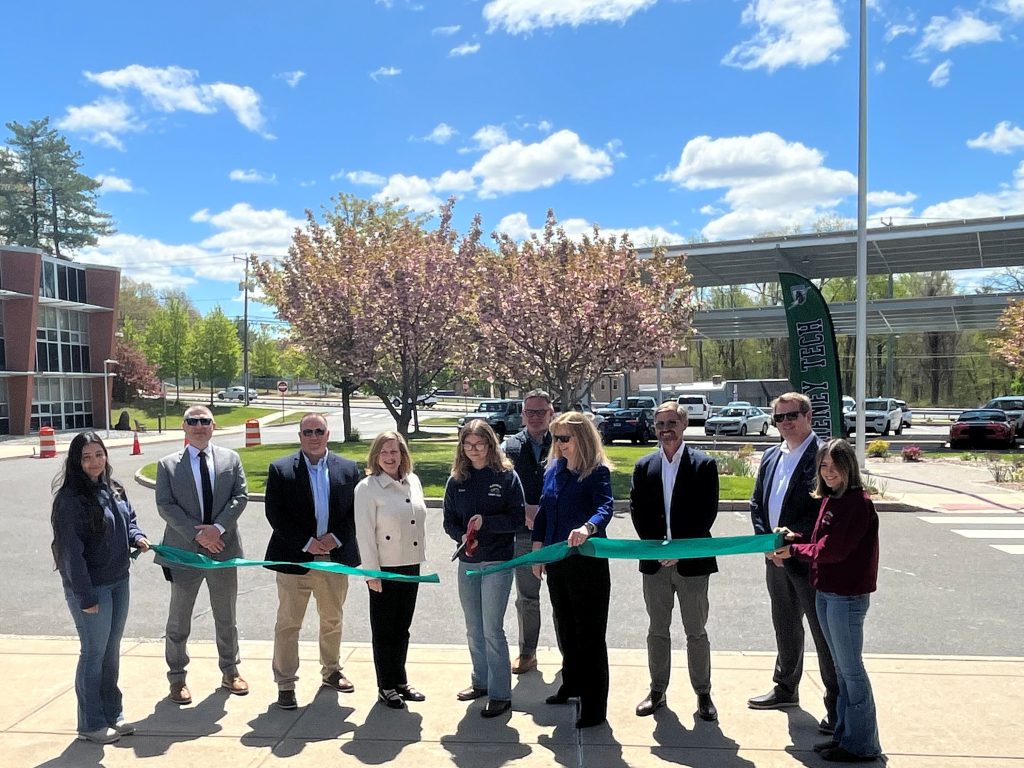 Students from Howell Cheney Technical High School cut the ribbon on the new carport solar energy system (in background), joined by Principal David Batch, DAS Commissioner Michelle Gilman, CTECS Executive Director Dr. Alice Pritchard, and project partners from the Connecticut Green Bank and Verogy.